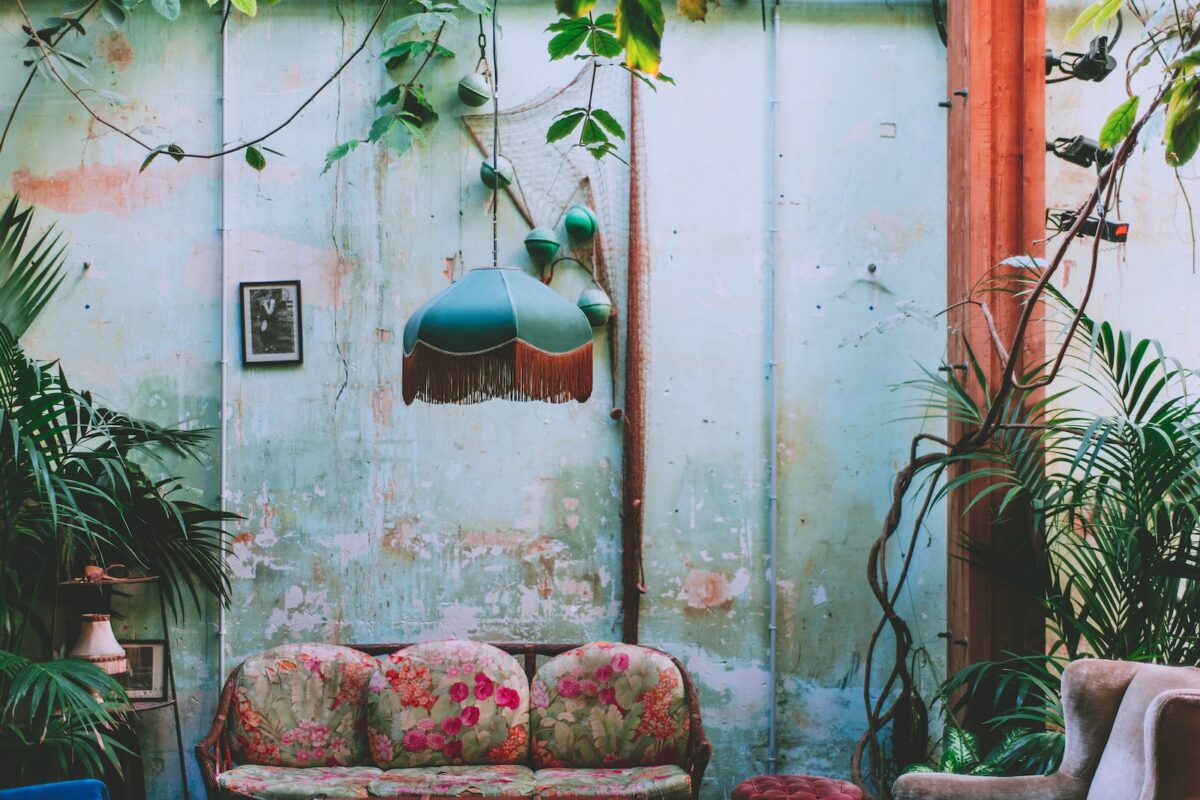 Interior of room with vintage chandelier and green plants growing above old coach at shabby wall with wooden column